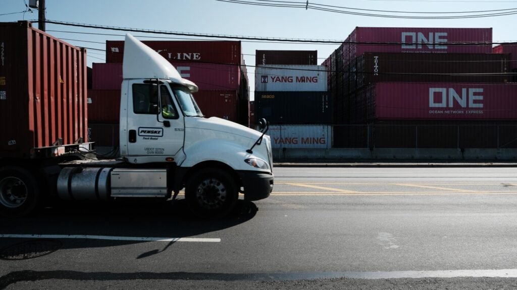 Diesel drayage truck hauling stacked shipping containers past a row of port cargo boxes